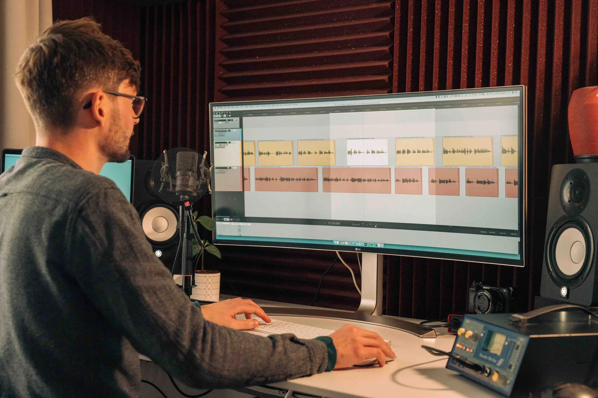 Stemacteur bewerkt voice-overtracks in zijn homestudio, microfoon naast het scherm.
